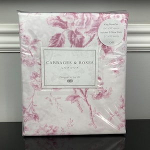 May include: A king duvet set in a clear plastic package. The bedding features a white background with a pink floral pattern. The packaging includes a label with the text "Cabbages & Roses London" and "Designed in the UK".