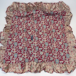 Op de afbeelding: Een rood, cr&egrave;me en blauw bloemenpatroon textiel met een gefranjerde rand. De stof heeft een herhalend bloemenontwerp in cr&egrave;me en blauw op een rode achtergrond. De randen zijn afgewerkt met een cr&egrave;mekleurige franje.