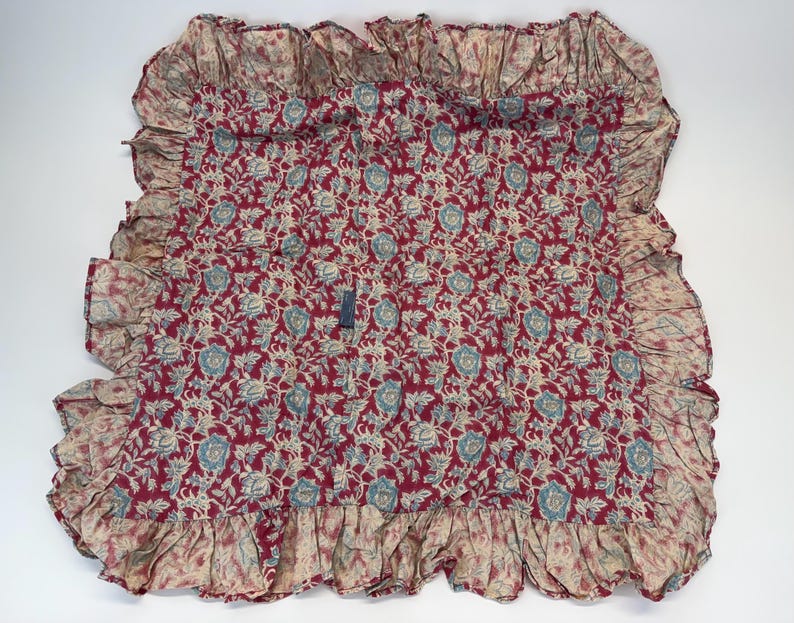 Op de afbeelding: Een rood, cr&egrave;me en blauw bloemenpatroon textiel met een gefranjerde rand. De stof heeft een herhalend bloemenontwerp in cr&egrave;me en blauw op een rode achtergrond. De randen zijn afgewerkt met een cr&egrave;mekleurige franje.