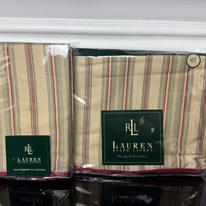 May include: A Lauren Ralph Lauren bedding set in clear plastic packaging, including two standard pillowcases and one queen flat sheet. The fabric features a vertical striped pattern in tan, sage green, and deep red, with dark green brand labels.