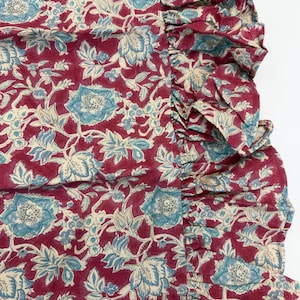 Op de afbeelding: Close-up van een stof met een bloemenpatroon. De stof heeft een dieprode achtergrond met een herhalend ontwerp van blauwe en cr&egrave;mekleurige bloemen en bladeren. Een gefranjerde rand is zichtbaar aan de rechterkant van de stof.