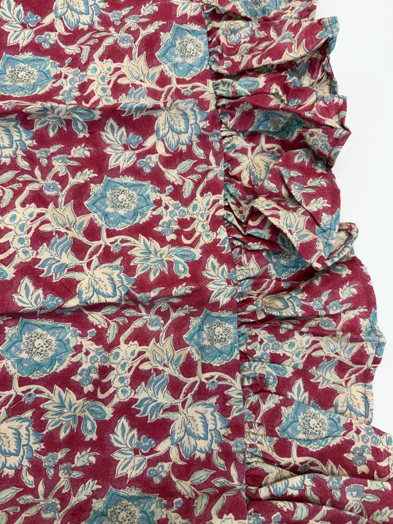 Op de afbeelding: Close-up van een stof met een bloemenpatroon. De stof heeft een dieprode achtergrond met een herhalend ontwerp van blauwe en cr&egrave;mekleurige bloemen en bladeren. Een gefranjerde rand is zichtbaar aan de rechterkant van de stof.
