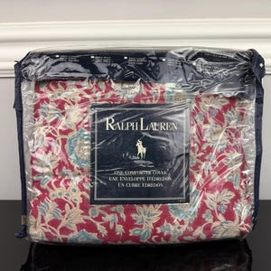 Op de afbeelding: Een verpakte Ralph Lauren dekbedovertrek. De overtrek heeft een bloemenpatroon in rood, blauw en beige. De verpakking bevat een marineblauw label met het Ralph Lauren logo en tekst in het Engels en Frans.