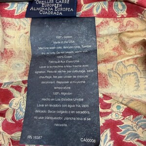 Op de afbeelding: Close-up van een kledinglabel met tekst in meerdere talen, waaronder "EUROPEAN SQUARE" en "100% Cotton". Het label is bevestigd aan een rood en cr&egrave;mekleurig bloemmotief. Wasvoorschriften zijn ook zichtbaar.