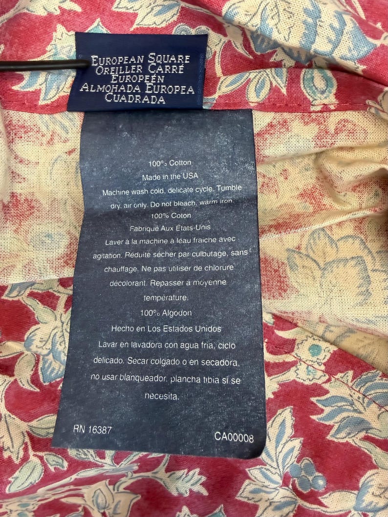 Op de afbeelding: Close-up van een kledinglabel met tekst in meerdere talen, waaronder "EUROPEAN SQUARE" en "100% Cotton". Het label is bevestigd aan een rood en cr&egrave;mekleurig bloemmotief. Wasvoorschriften zijn ook zichtbaar.