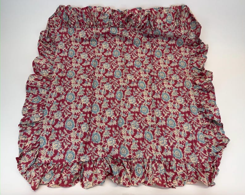 Op de afbeelding: Een bloemmotiefstof in tinten bordeauxrood, cr&egrave;me en blauwgroen. De stof heeft een herhalend patroon van bloemen en bladeren. De randen zijn afgewerkt met een gefranjerde rand. De stof wordt waarschijnlijk gebruikt voor woondecoratie.
