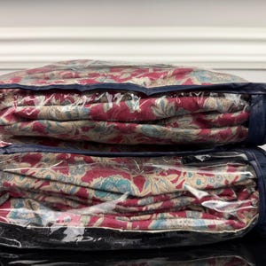 Op de afbeelding: Twee dekbedden met bloemenpatroon in doorzichtige opbergtassen met ritssluiting. De dekbedden hebben een rood, beige en turquoise bloemenontwerp. De tassen hebben marineblauwe randen en ritsen, gestapeld.