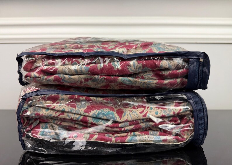 Op de afbeelding: Twee dekbedden met bloemenpatroon in doorzichtige opbergtassen met ritssluiting. De dekbedden hebben een rood, beige en turquoise bloemenontwerp. De tassen hebben marineblauwe randen en ritsen, gestapeld.