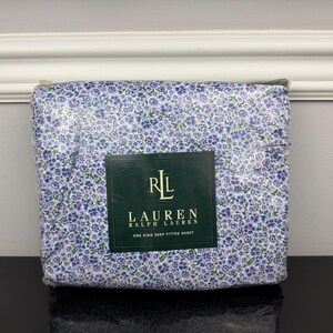 May include: A folded king-size deep fitted sheet set. The sheet set features a floral pattern in shades of blue, purple, and white. The packaging includes a dark green label with the brand logo and text.