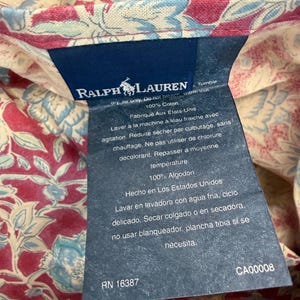 Op de afbeelding: Een marineblauw Ralph Lauren-label met witte tekst en een logo van een persoon op een paard. Het label is bevestigd aan een bloemenstof met rode, blauwe en cr&egrave;metinten. Het label bevat wasvoorschriften.