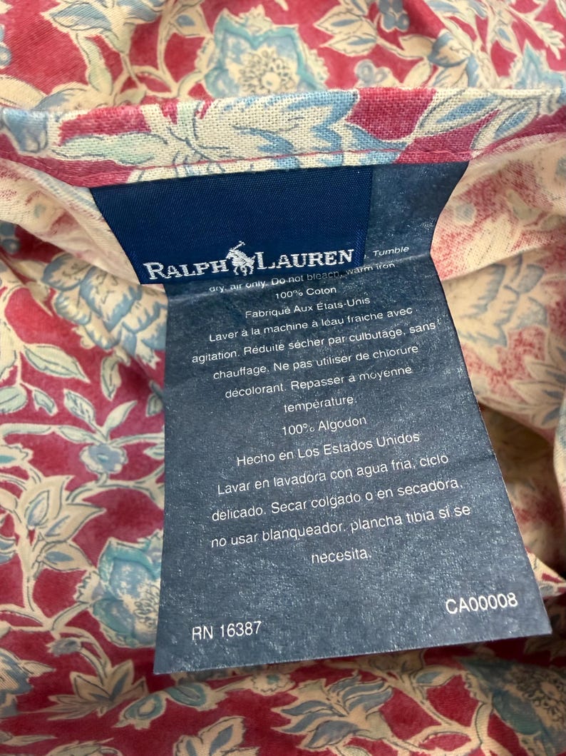 Op de afbeelding: Een marineblauw Ralph Lauren-label met witte tekst en een logo van een persoon op een paard. Het label is bevestigd aan een bloemenstof met rode, blauwe en cr&egrave;metinten. Het label bevat wasvoorschriften.