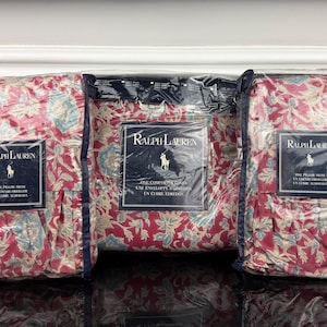 Op de afbeelding: Drie verpakte kussenslopen met een bloemenpatroon in rood, blauw en cr&egrave;me. Elk pakket heeft een label met het Ralph Lauren-logo en tekst. De kussenslopen zitten in een transparante plastic verpakking.
