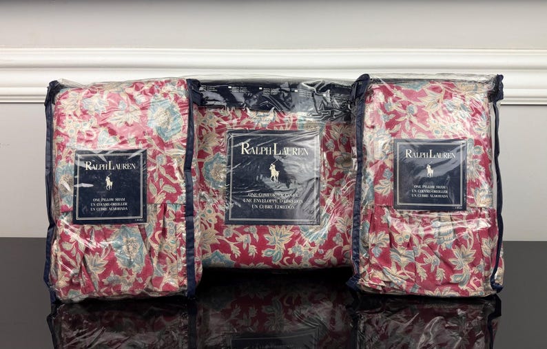 Op de afbeelding: Drie verpakte kussenslopen met een bloemenpatroon in rood, blauw en cr&egrave;me. Elk pakket heeft een label met het Ralph Lauren-logo en tekst. De kussenslopen zitten in een transparante plastic verpakking.