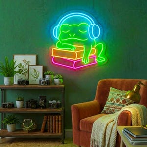 Puede incluir: Un letrero de neón que muestra una rana verde brillante con auriculares azules, descansando sobre una pila de dos libros. La rana aparece con los ojos cerrados y una postura relajada, emitiendo un brillo vibrante sobre la pared.
