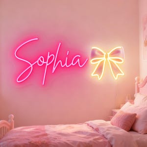May include: A neon sign in a bedroom spells out "Sophia" in bright pink script, accompanied by a yellow bow-shaped light. The room is decorated in shades of pink, with a bed and pillows visible.
