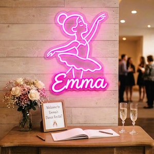 Op de afbeelding: Een felroze neonbord met de omtrek van een ballerina in een tutu met de naam Emma in cursief schrift eronder. Het bord is bevestigd aan een lichtgekleurde houten wand boven een tafel met een boeket bloemen en een bordje.
