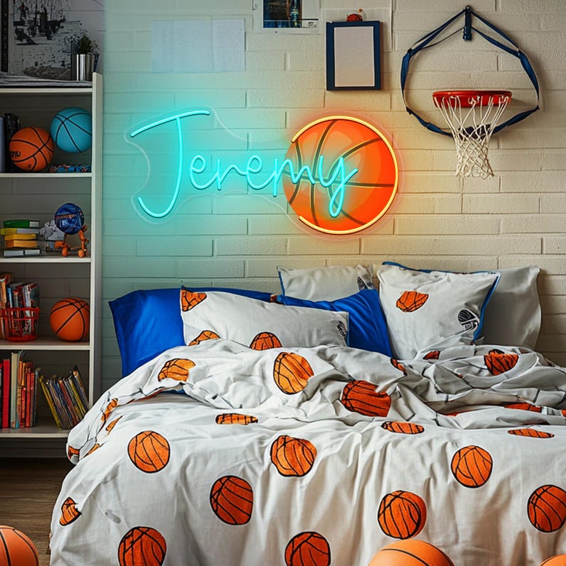 May include: A white bed with a basketball themed duvet cover. The duvet cover has a white background with orange basketballs printed on it. The bed has blue and white pillows. A neon sign with the name "Jeremy" and a basketball is on the wall behind the bed.