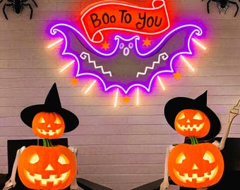 Op de afbeelding: Halloween-thema decoraties. Een neonbord in de vorm van een vleermuis met de tekst "Boo To You" op een banner. Twee pompoenhoofdige skeletten met zwarte heksenhoeden zitten op stoelen. Zwarte spinnen decoraties.