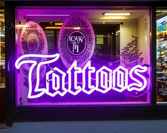 May include: A neon sign in a shop window spells out "Tattoos" in a bold, white font, illuminated by a vibrant purple glow. The shop window also features decorative oval-shaped panels with intricate black designs.