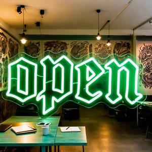 以下が含まれることがあります： 大胆な書体で「OPEN」と書かれた明るい緑色のネオンサイン。透明な裏板に取り付けられ、タトゥーショップに吊り下げられています。前景にはテーブルと椅子があり、壁にはタトゥーアートが飾られています。