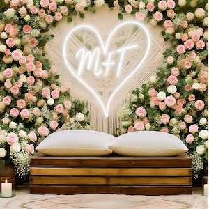 May include: A wedding backdrop featuring a large neon heart with the letters "M+T" inside, surrounded by pink and white roses and greenery. Two white pillows rest on a wooden crate, creating a romantic setting.