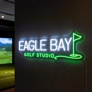 Op de afbeelding: Een aan de muur gemonteerd neonbord met de tekst EAGLE BAY GOLF STUDIO in helder wit en groen licht. Het ontwerp bevat een wit golfvlag-icoon aan de rechterkant, met het woord EAGLE BAY in het wit en GOLF STUDIO in het groen eronder.