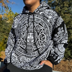 May include: Black and white hooded sweatshirt with an intricate Aztec-inspired design featuring a central face and geometric patterns.