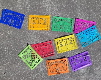 May include: A colorful papel picado banner with cut-out designs. The banner features rectangular flags in various colors, including blue, yellow, green, pink, red, orange, and purple. The designs include skulls and other decorative patterns, strung together on a string.