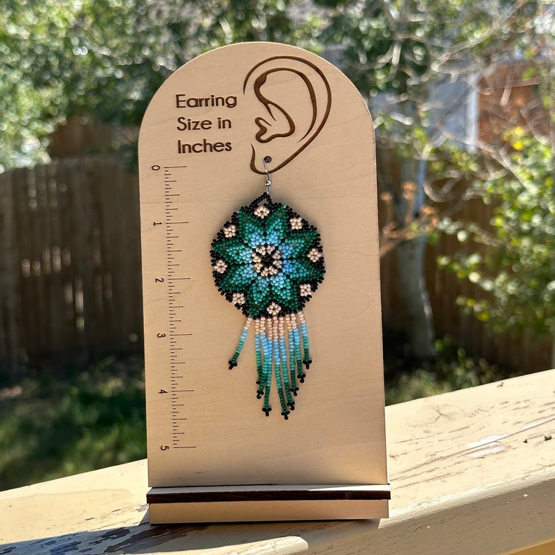 Op de afbeelding: Groene, blauwe en witte kralen oorring met een bloemenpatroon en franjes. De oorring wordt getoond op een houten oorringstandaard met een liniaal die aangeeft dat de oorring ongeveer 7,6 cm lang is. "Earring Size in Inches" staat op de standaard.