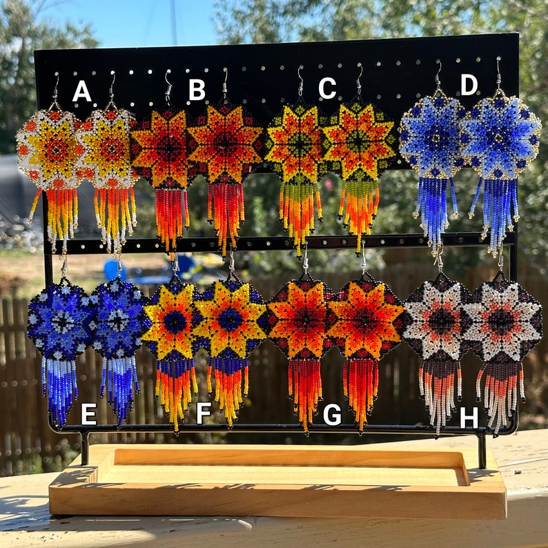 Op de afbeelding: Acht paar geperde oorbellen op een displaystandaard. De oorbellen zijn gemaakt met kleurrijke kralen in ster- en bloemenpatronen. De oorbellen zijn van A tot en met H gelabeld.