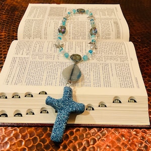 Puede incluir: Un colgante de cruz azul y blanco con un collar de cuentas con detalles azules y plateados. El collar está sobre una Biblia abierta.
