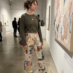 May include: A long-sleeved olive green crewneck shirt paired with patchwork trousers featuring a variety of printed fabric squares in shades of beige, cream, black, and muted pink. The outfit includes a brown leather crossbody bag and dark boots.