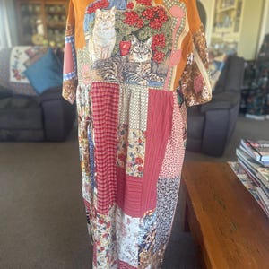 May include: A long-sleeved dress with a mustard yellow top featuring a graphic of two cats sitting among red flowers. The lower portion of the dress is constructed from a patchwork of various fabrics in shades of red, white, and brown with floral and plaid patterns.