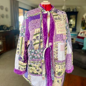 May include: A patchwork jacket featuring various floral and polka dot patterns in shades of purple, lavender, and cream. The garment includes a bright purple faux fur trim along the front opening and cuffs, with a tie closure and visible text.
