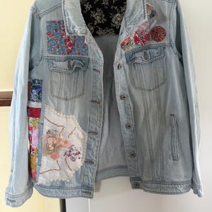 Peut inclure: Une veste en jean bleu clair ornée de divers empiècements en tissu, dont un carré à fleurs bleues, une pièce en dentelle blanche brodée et plusieurs rosettes en tissu circulaires sur la poitrine. Le col intérieur est doublé de tissu noir à fleurs.