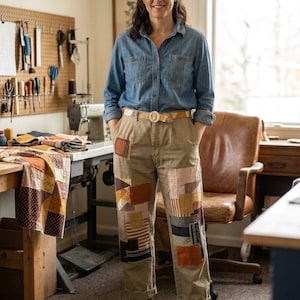 May include: A blue denim button-down shirt with long sleeves and tan trousers featuring various rectangular fabric patches in shades of brown, orange, and yellow. The trousers have a light-coloured belt with a circular buckle and are cuffed at the ankles.