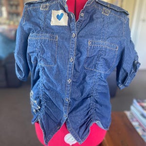 May include: A dark blue denim button-down shirt featuring a mandarin collar, shoulder epaulets, and two chest pockets. The front displays a white fabric patch with a blue heart graphic. The sides have vertical ruched detailing and short sleeves.