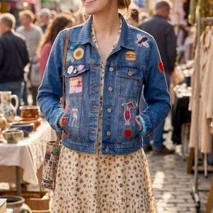 May include: A blue denim jacket featuring various colourful embroidered patches, including a bee, a burger, a cocktail, and a sunflower, worn over a cream-coloured maxi dress with a small brown floral print and a tiered skirt design.