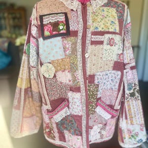 May include: A long-sleeved jacket constructed from a patchwork of various floral and patterned fabrics in shades of pink, brown, and cream. The garment features a white lace collar, front button closures, and two front pockets trimmed with lace.