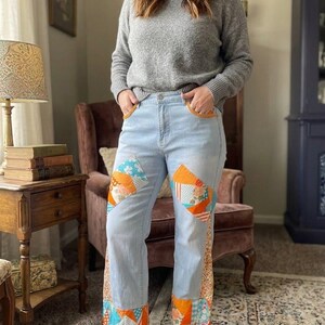May include: Light blue denim jeans featuring patchwork panels with orange, white, and blue floral and geometric patterns on the thighs and cuffs, along with orange crocheted trim on the front pockets, paired with a grey long-sleeve sweater.