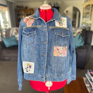 May include: A blue denim jacket featuring a collar, long sleeves, and button-down front. The jacket is decorated with various rectangular and floral-shaped fabric patches, including images of wine bottles, glasses, and grapes, stitched onto the front.