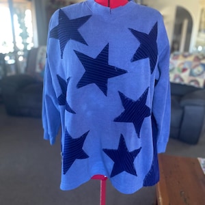 May include: A periwinkle blue long-sleeve sweatshirt featuring a repeating pattern of dark navy blue stars. The stars have a subtle horizontal striped texture and frayed edges. The garment has a crew neckline and ribbed cuffs on the sleeves.