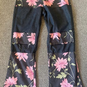 May include: Dark charcoal denim trousers featuring a floral pattern with pink flowers and green leaves. The trousers have a high-waisted floral section, solid dark denim mid-sections, rectangular floral patches on the knees, and floral flared leg bottoms.