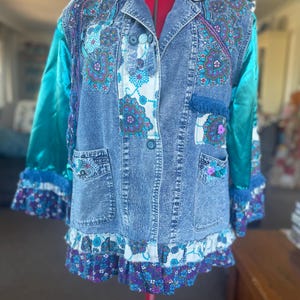 May include: A blue acid-wash denim jacket featuring teal satin sleeves, floral patterned fabric panels, and ruffled trim at the hem and cuffs. The jacket includes button closures, two front pockets, and decorative embellishments like small buttons.