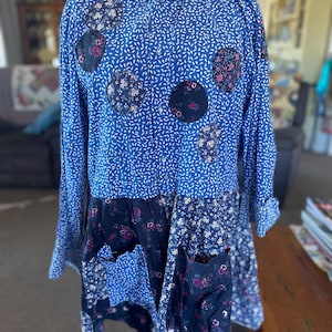 May include: A long-sleeved button-down shirt featuring a blue base with a white leaf-like pattern. The garment is accented with dark navy circular patches containing floral designs and includes two large front pockets with mixed floral patterns.