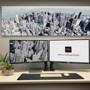 May include: A modern office setup featuring two computer monitors displaying cityscapes and the MementoDesignStudio logo. A panoramic cityscape artwork hangs above the monitors. A keyboard, mouse, and potted plants complete the desk arrangement.