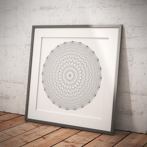Puede incluir: Un diseño geométrico en blanco y negro en forma circular está enmarcado en un marco plateado. El marco está sobre un suelo de madera.