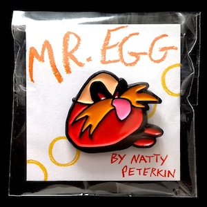 Op de afbeelding: Een rode en oranje emaille pin met een cartoonfiguur met een snor en de tekst "MR. EGG BY NATTY PETERKIN".