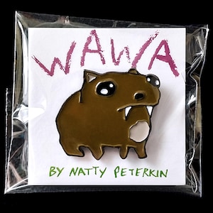 Puede incluir: Un pin de esmalte marrón y blanco con un capibara de dibujos animados con ojos grandes y dientes. El pin está empaquetado en una bolsa de plástico transparente con una tarjeta blanca que dice "Wawa" en rosa y "By Natty Peterkin" en verde.