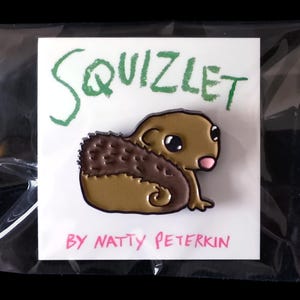 May include: A Squizlet enamel pin on a white card, packaged in a clear plastic sleeve. The pin features a cartoonish brown creature with a spiky back, large eyes, and a pink nose. The word "SQUIZLET" is written in green above the pin, and "BY NATTY PETERKIN" is written in pink below.